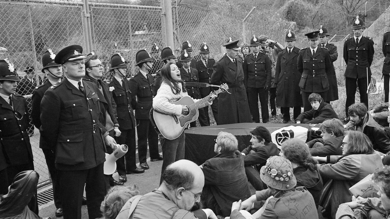 Police stand behind American Folk singer Julie Felix as she sings protest songs to marchers outside the gates of Regional Seat of Government No. 6 on April 13, 1968 in Berkshire, England.